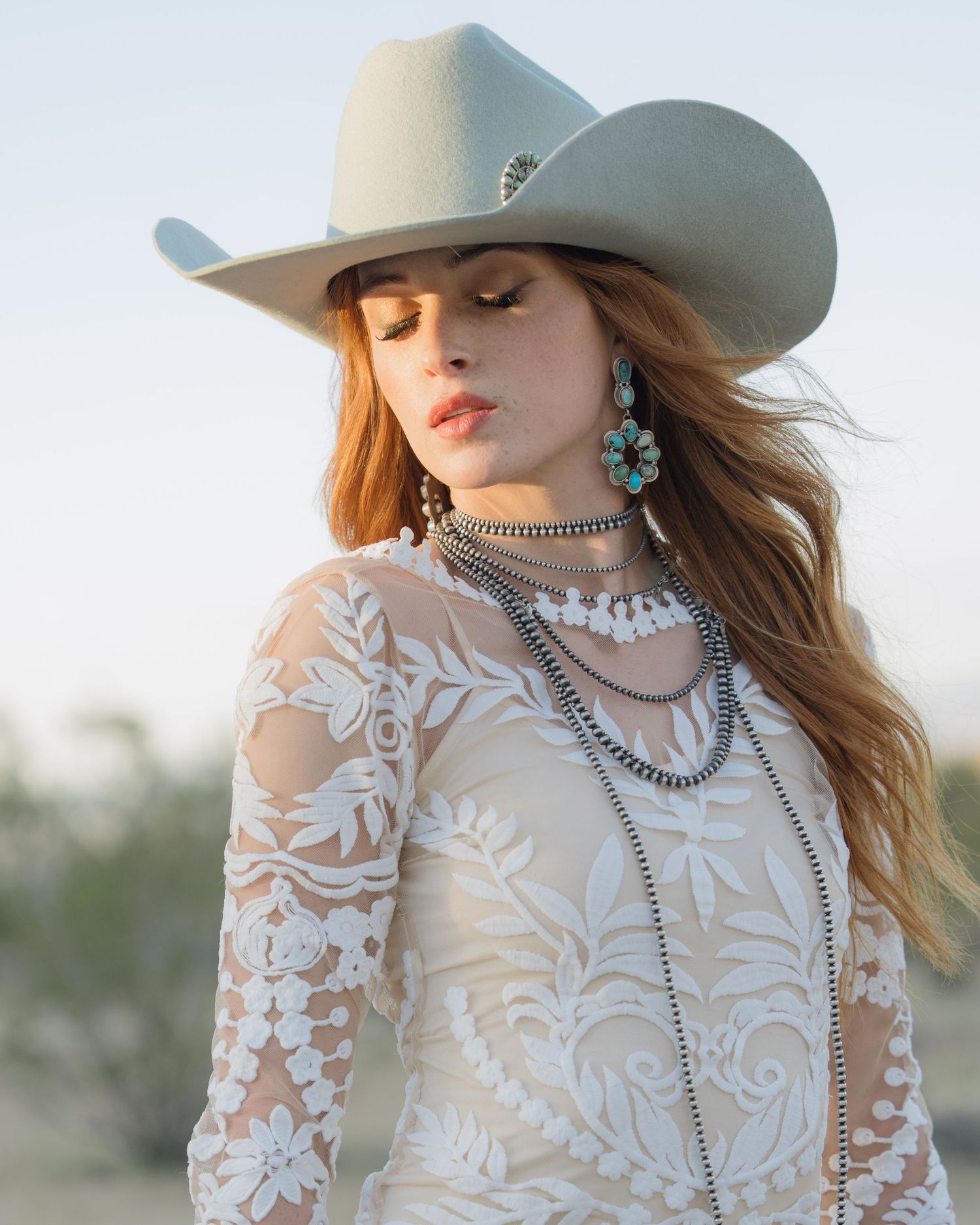 Elegant bride wearing lace dress, turquoise earrings, and gray cowboy hat in desert landscape.