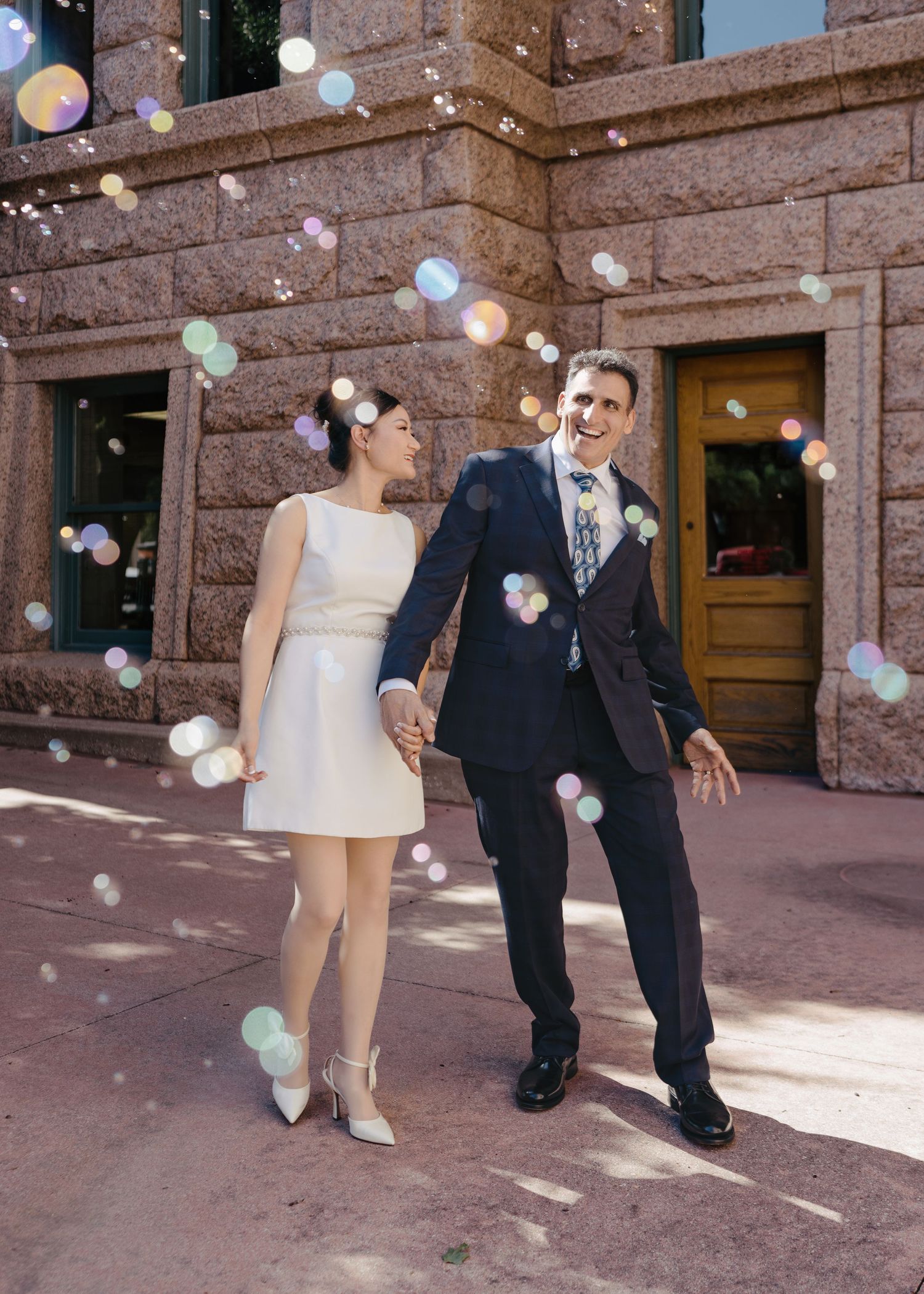 Newlywed couple holds hands surrounded by floating bubbles outside historic stone building.