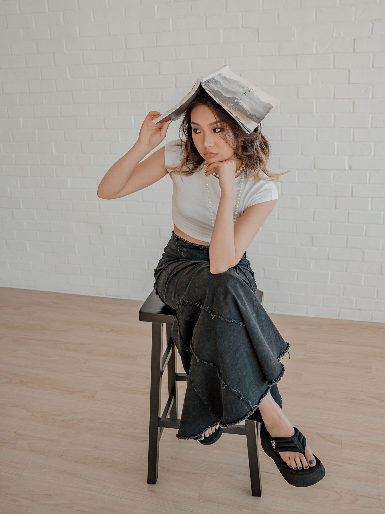 Young woman in white crop top balancing book on head while sitting on stool in bright studio.