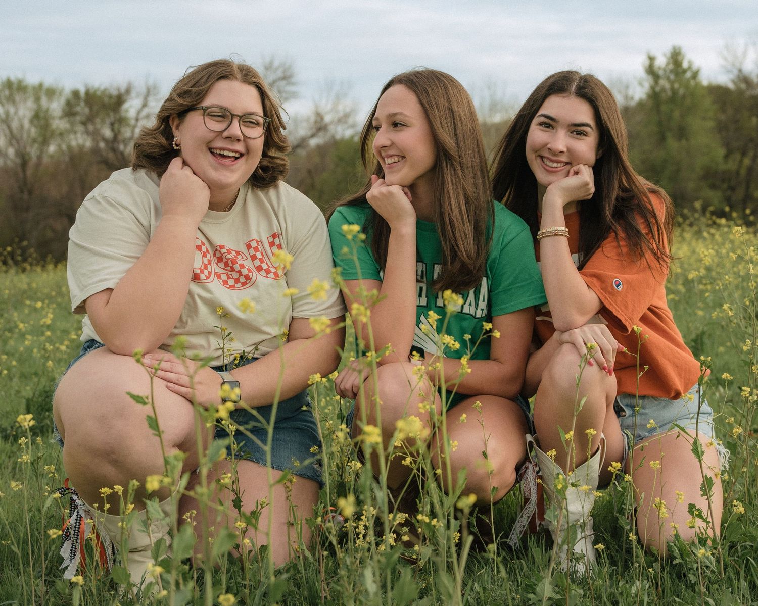 Three laughing girls crouching among yellow wildflowers wearing OSU and college graphic tees.