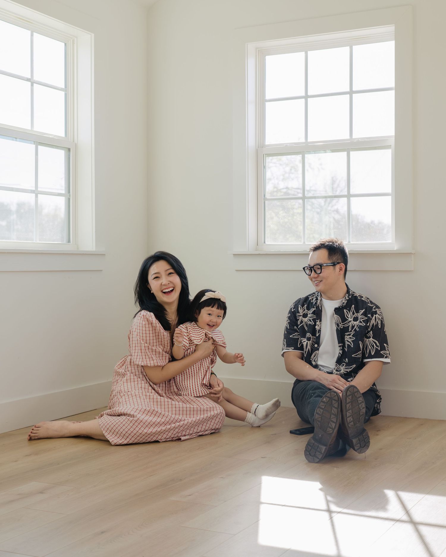 A laughing Asian family sitting on a sunny hardwood floor, mother and baby in matching pink striped outfits.