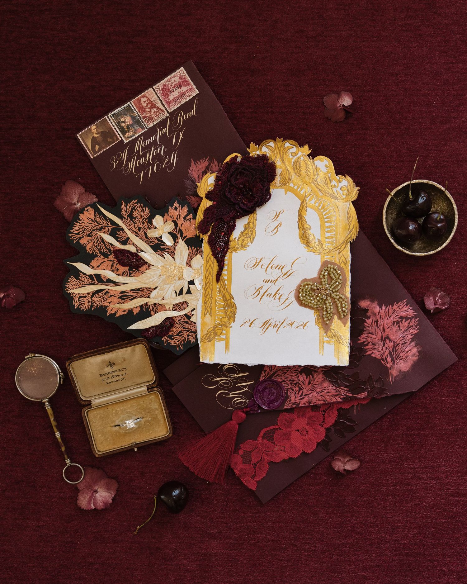 Ornate gold and burgundy wedding invitation suite with ring box and floral accents on red.