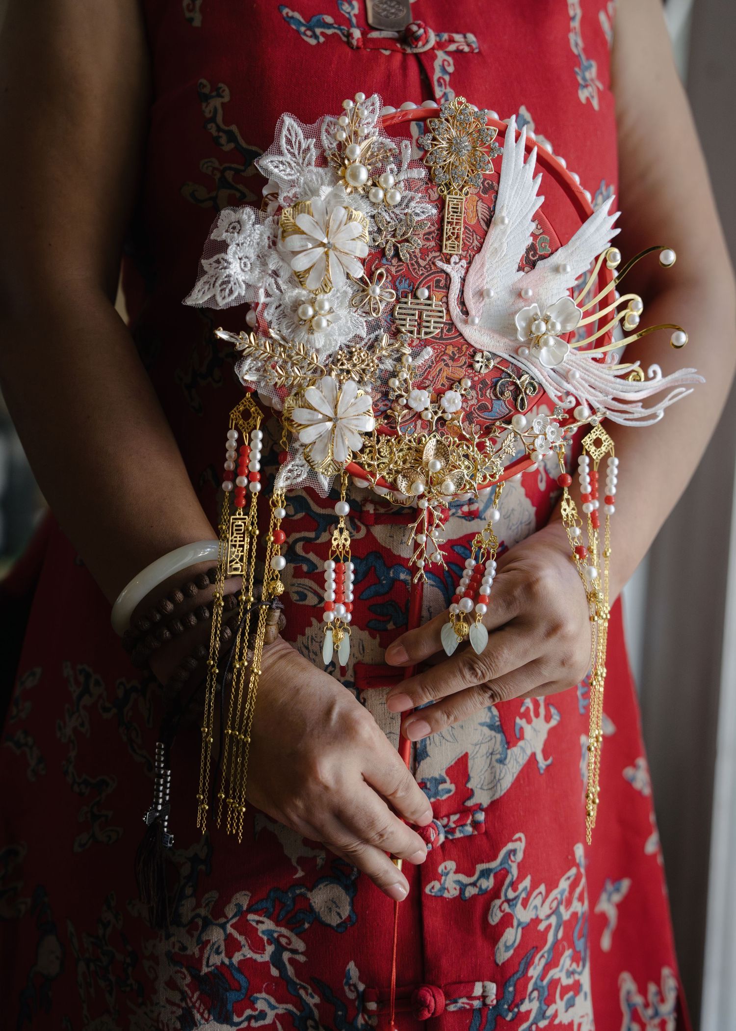 Close-up of ornate Chinese bridal fan with crane, pearls, and gold tassels held in red dress.