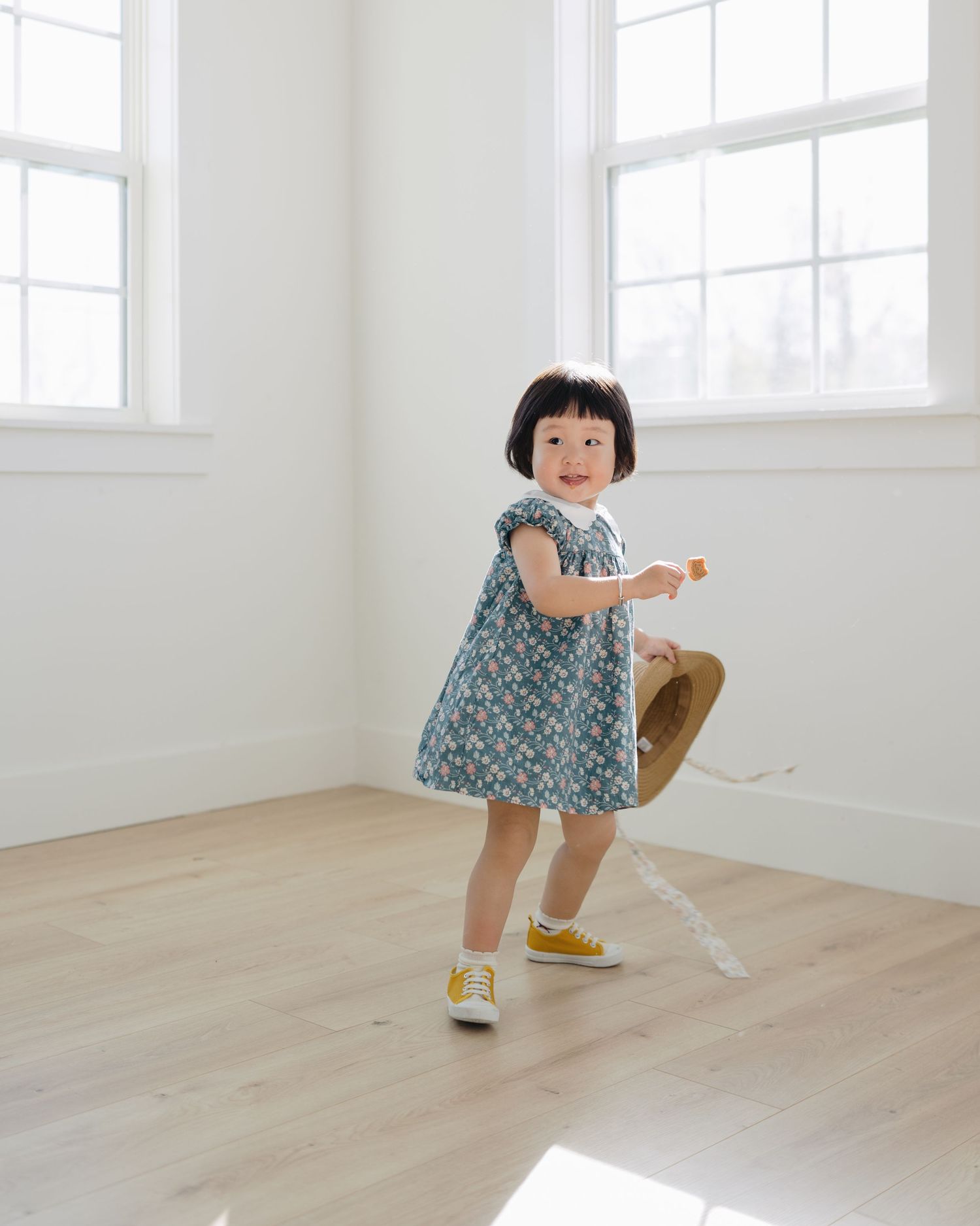 Adorable toddler girl in floral dress and yellow sneakers playing with straw hat indoors.