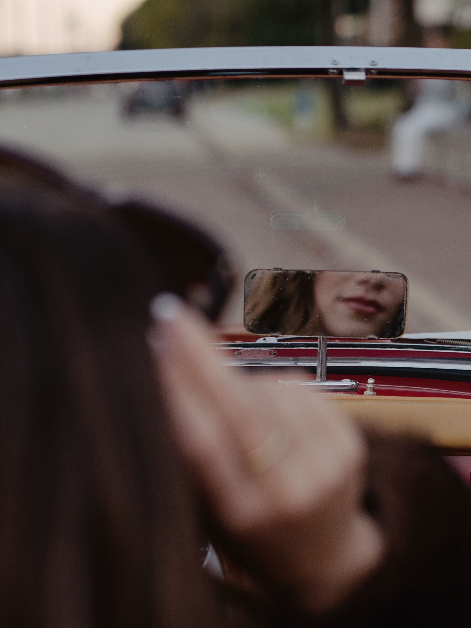 Woman's reflection visible in vintage red convertible car rearview mirror.