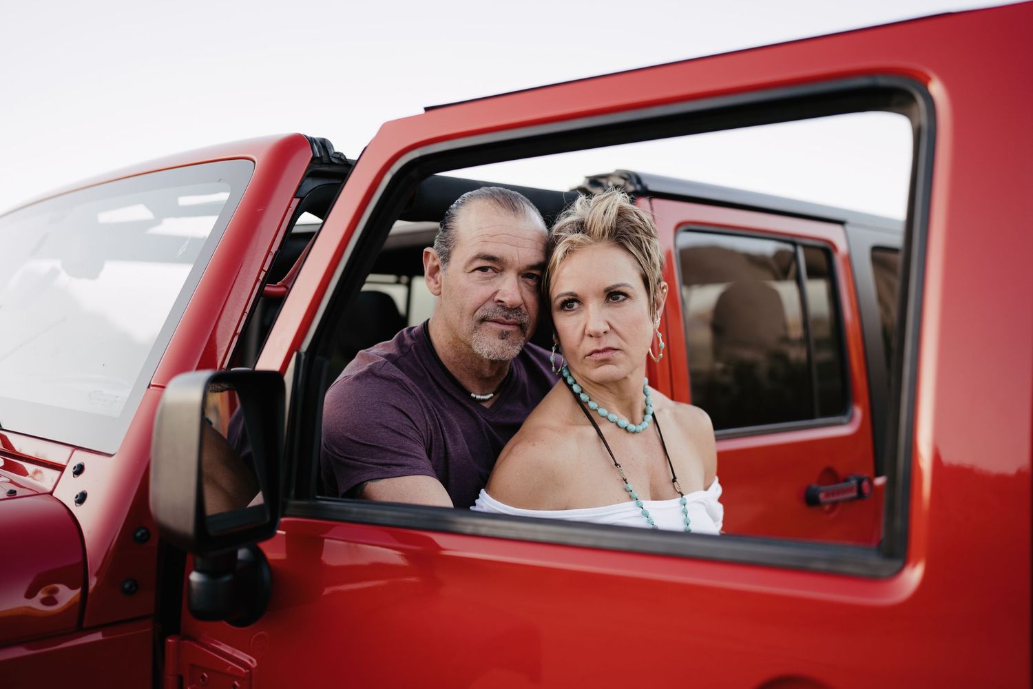 Man and woman posing together through the window of a red Jeep in an outdoor setting.