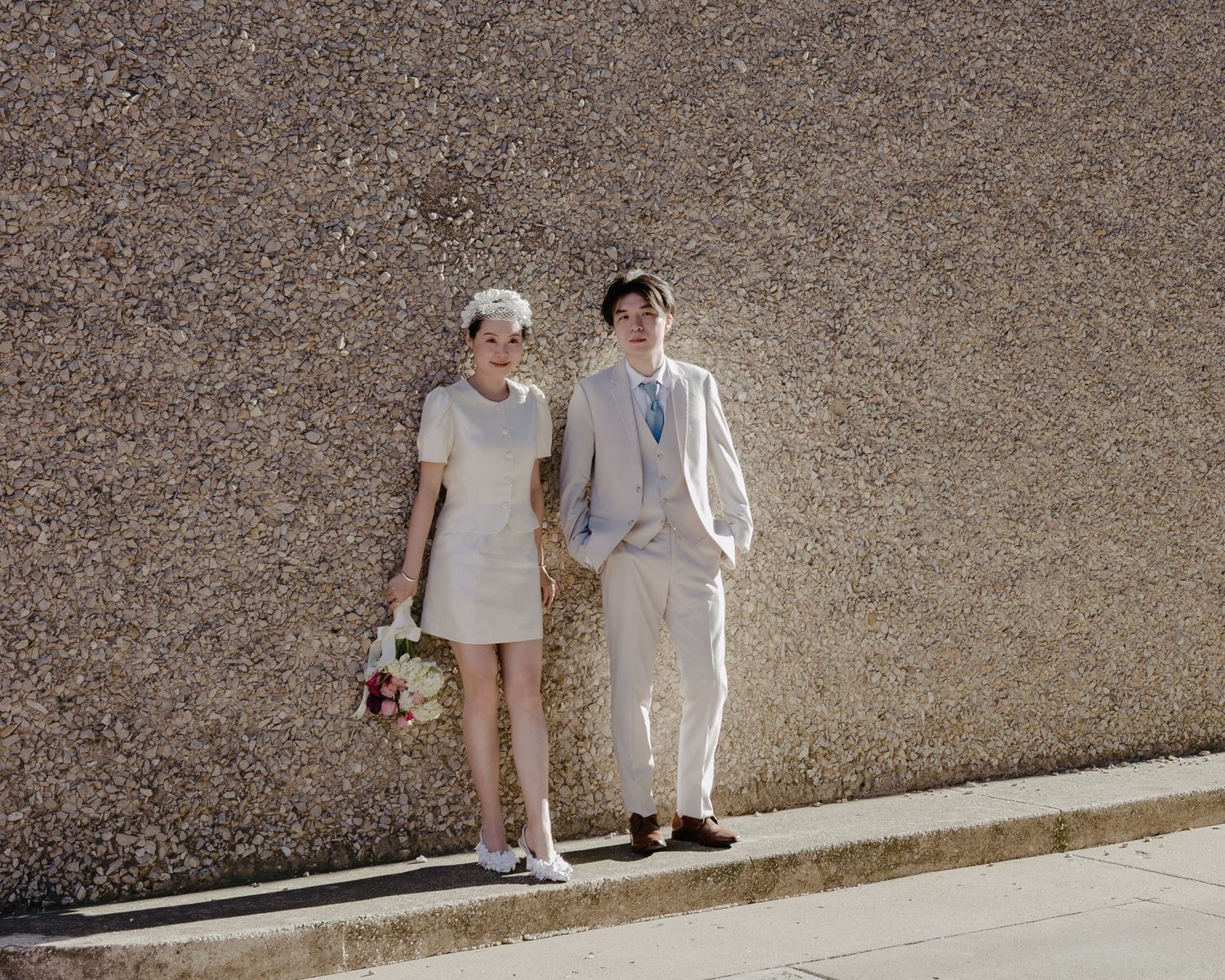 Bride in white mini dress and groom in cream suit pose together against a textured gray wall outdoors.