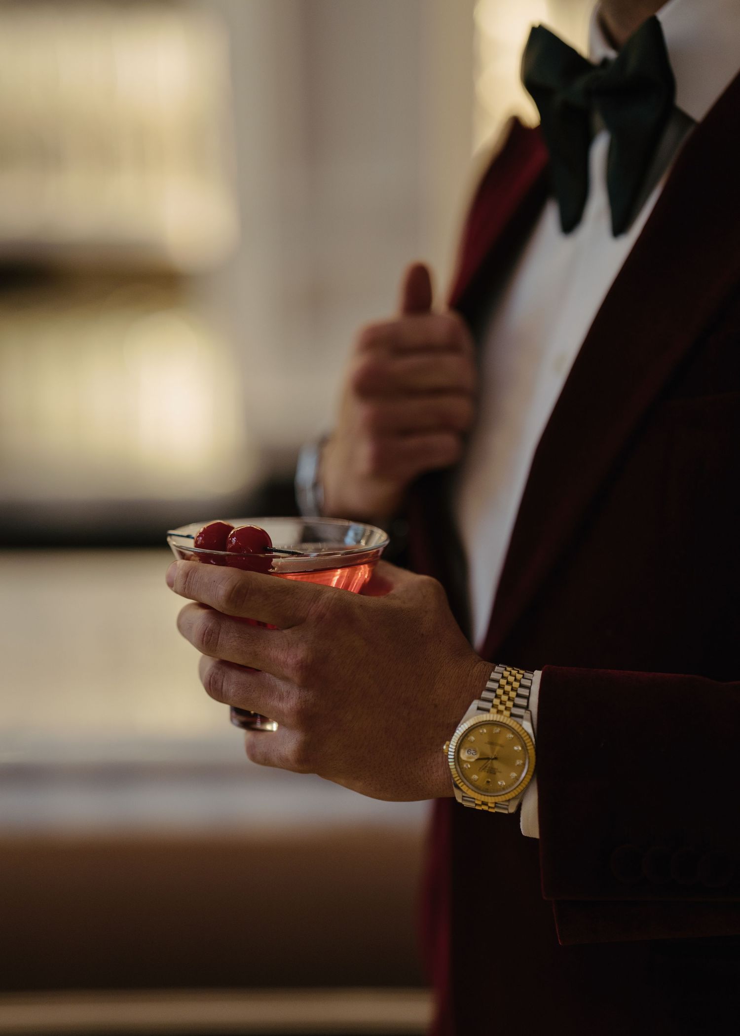 Groom in burgundy tuxedo holding cocktail glass with raspberries and gold Rolex watch.