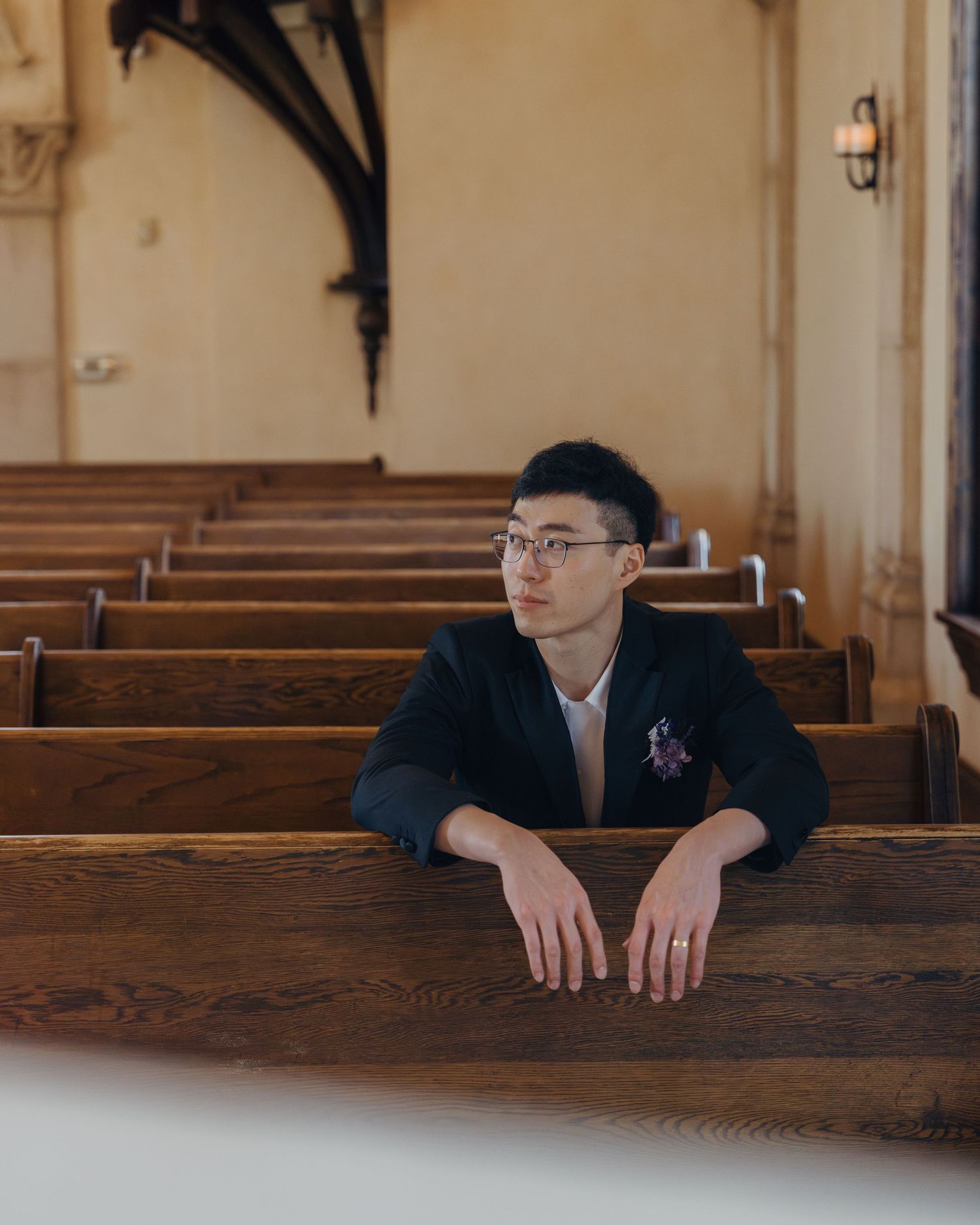 Young Asian man in black blazer with boutonniere resting arms on wooden church pew during wedding.