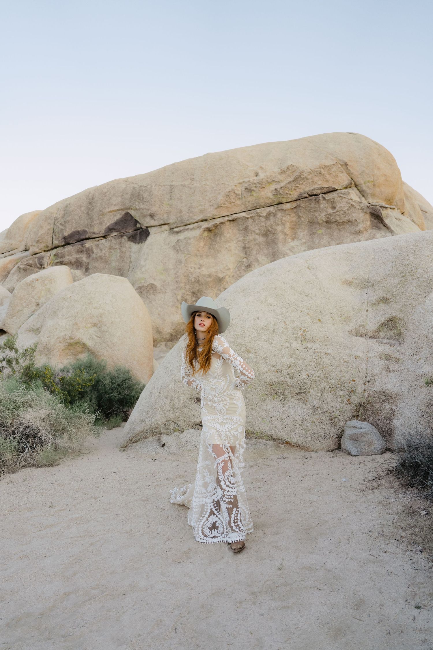 Boho bride in white lace maxi dress and gray hat posing near large desert boulders.