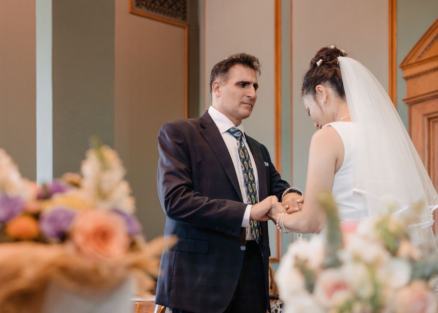 Groom placing ring on brides hand during intimate indoor wedding ceremony with floral decor.