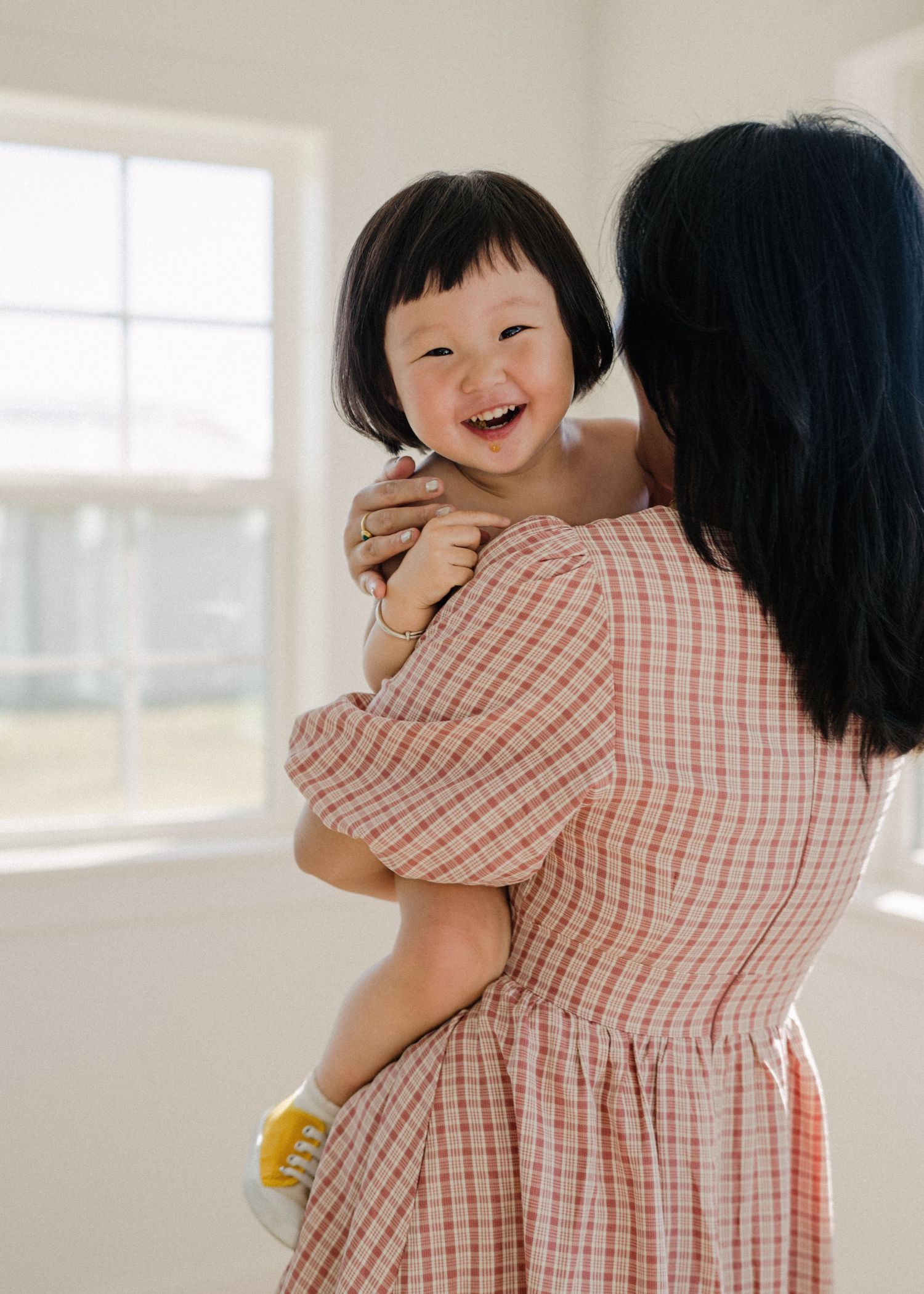 Joyful toddler girl laughing while being held by mother in pink plaid dress indoors.