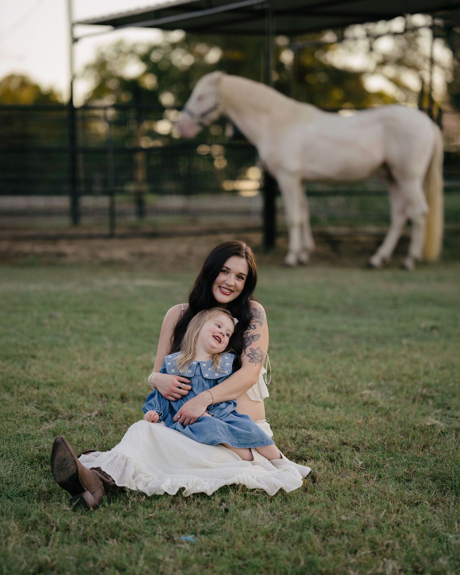 Mother hugging young daughter on grass at golden hour with white horse grazing in background.