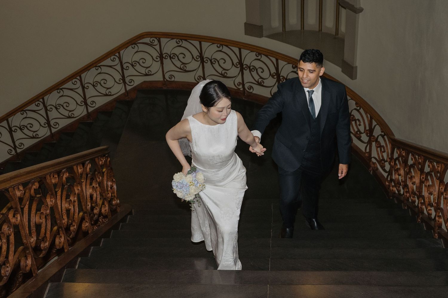 Bride and groom walking up elegant staircase with ornate railing, bride holding white bouquet in wedding dress.