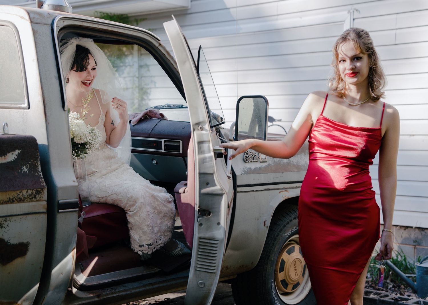 Bride in lace wedding dress exits vintage truck beside bridesmaid in red satin gown.