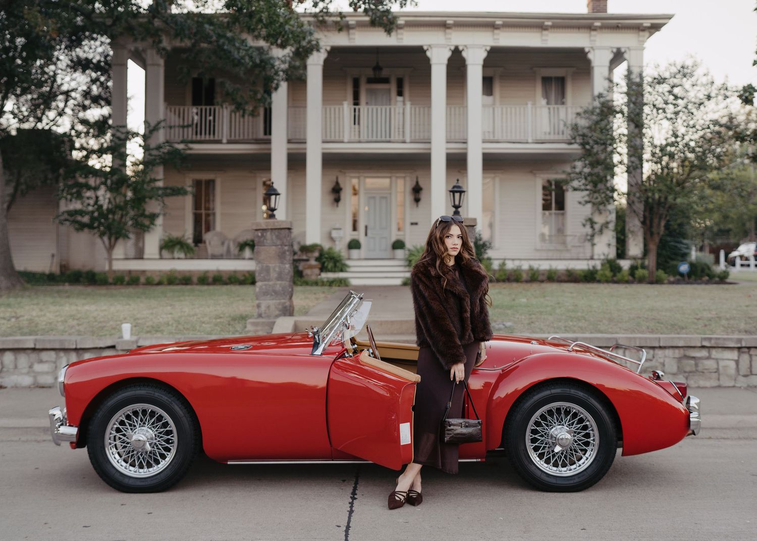 Stylish woman in fur coat standing beside a vintage red MG roadster in front of a historic home.
