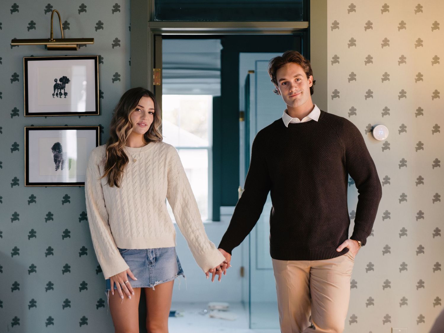 Stylish couple holding hands in a room with blue patterned wallpaper and framed wall art.