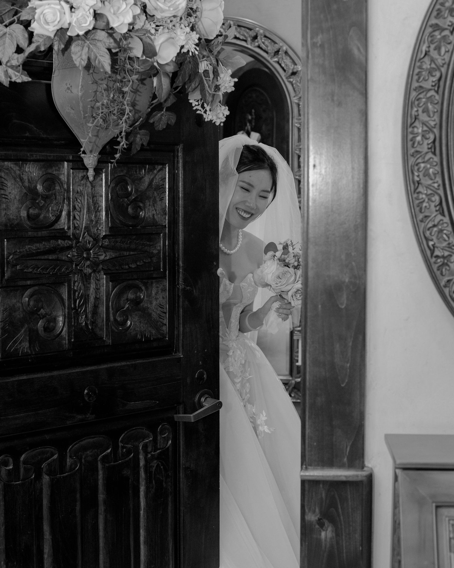 Black and white photo of a smiling bride peeking through an ornate church door with her bouquet.