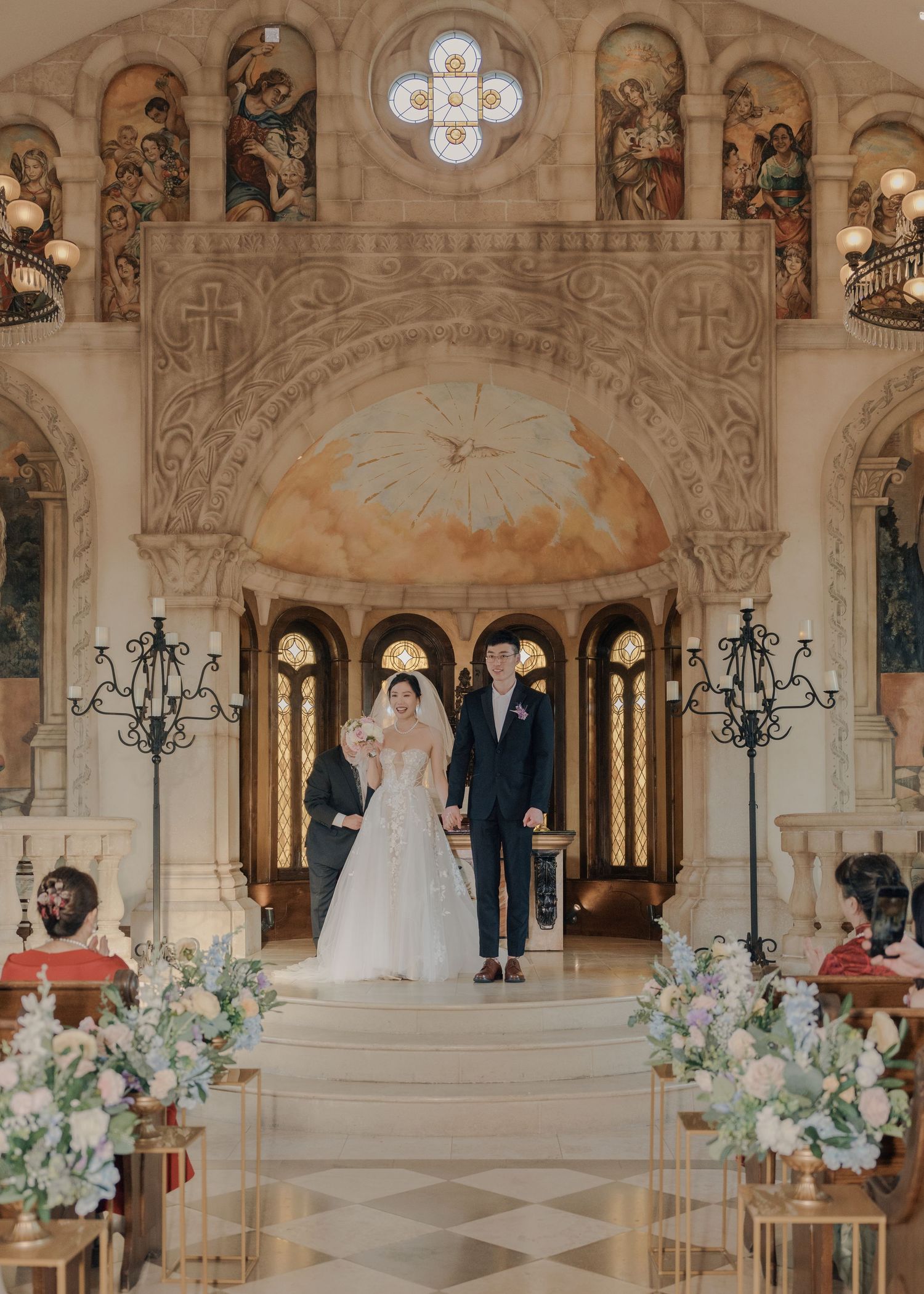Bride and groom walk down the aisle of an ornate chapel with floral arrangements and arched windows.