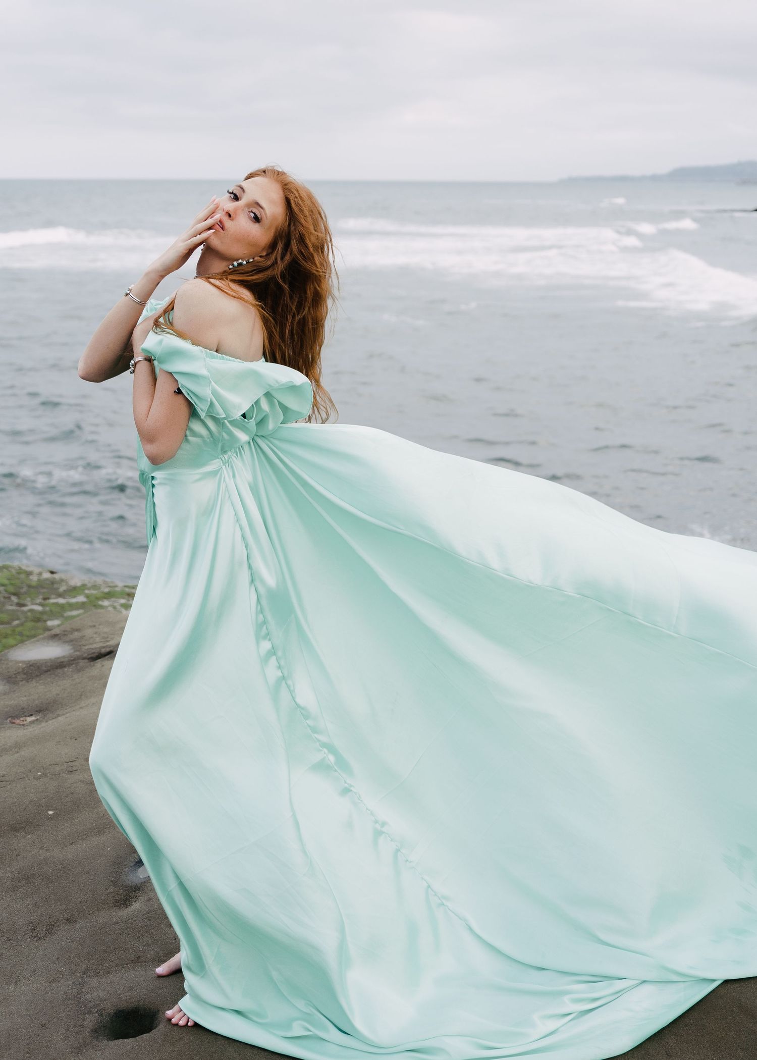 Redhead woman in flowing mint off-shoulder ball gown posing dramatically by the ocean.