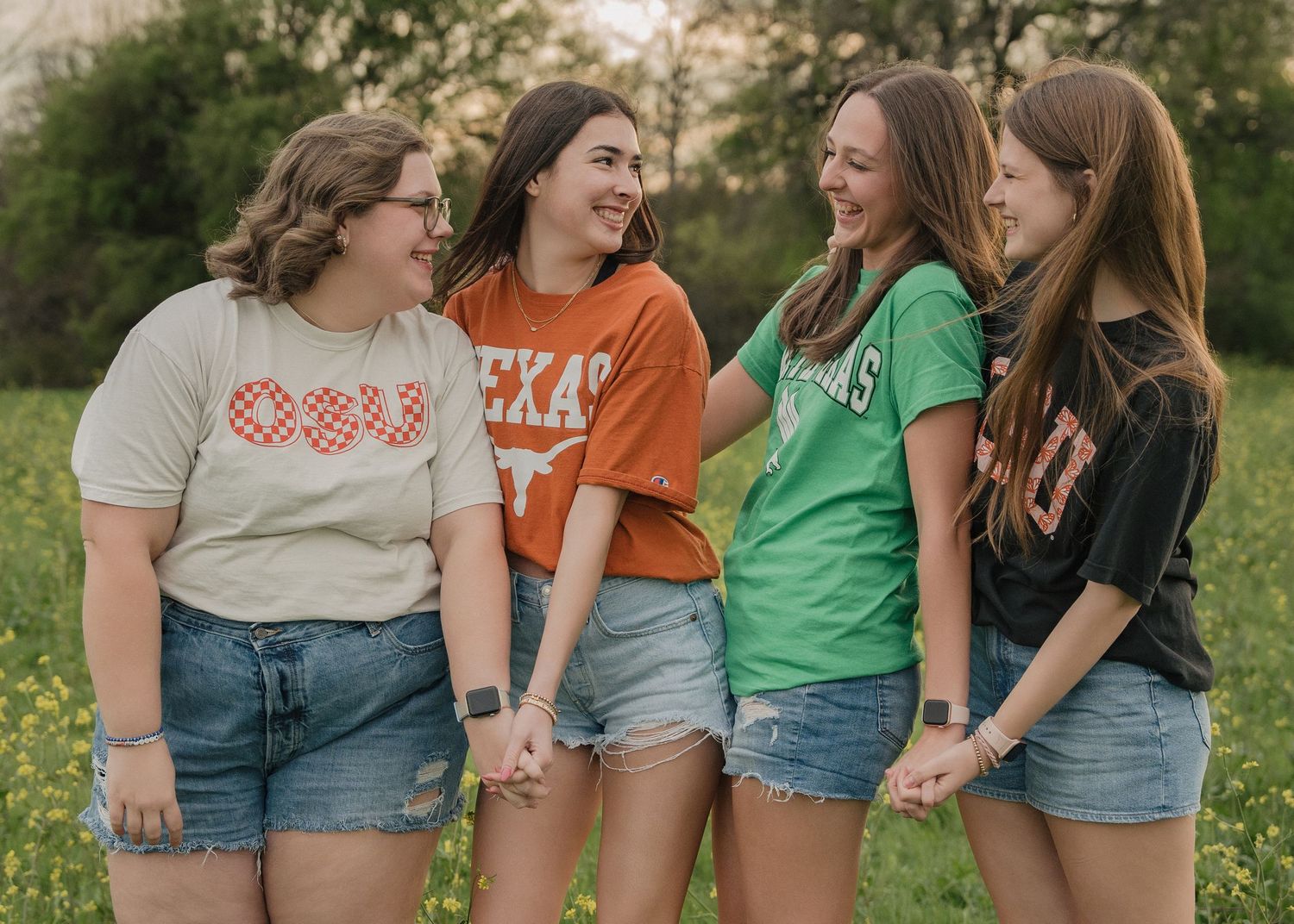 Four young women in college graphic tees hold hands and laugh together in an outdoor meadow setting.