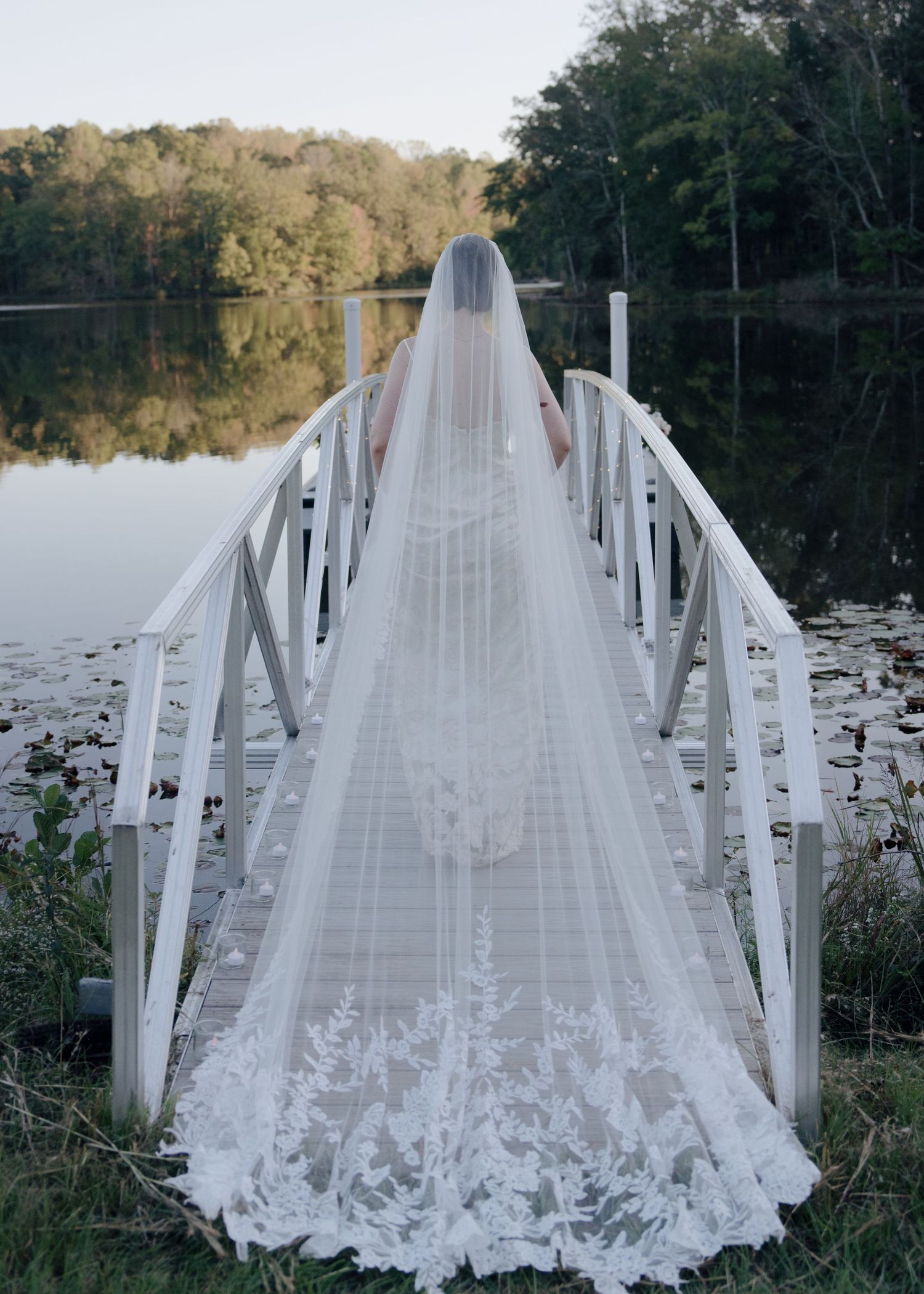 Bride in cathedral lace-trimmed veil standing on white wooden dock over serene autumn lake.