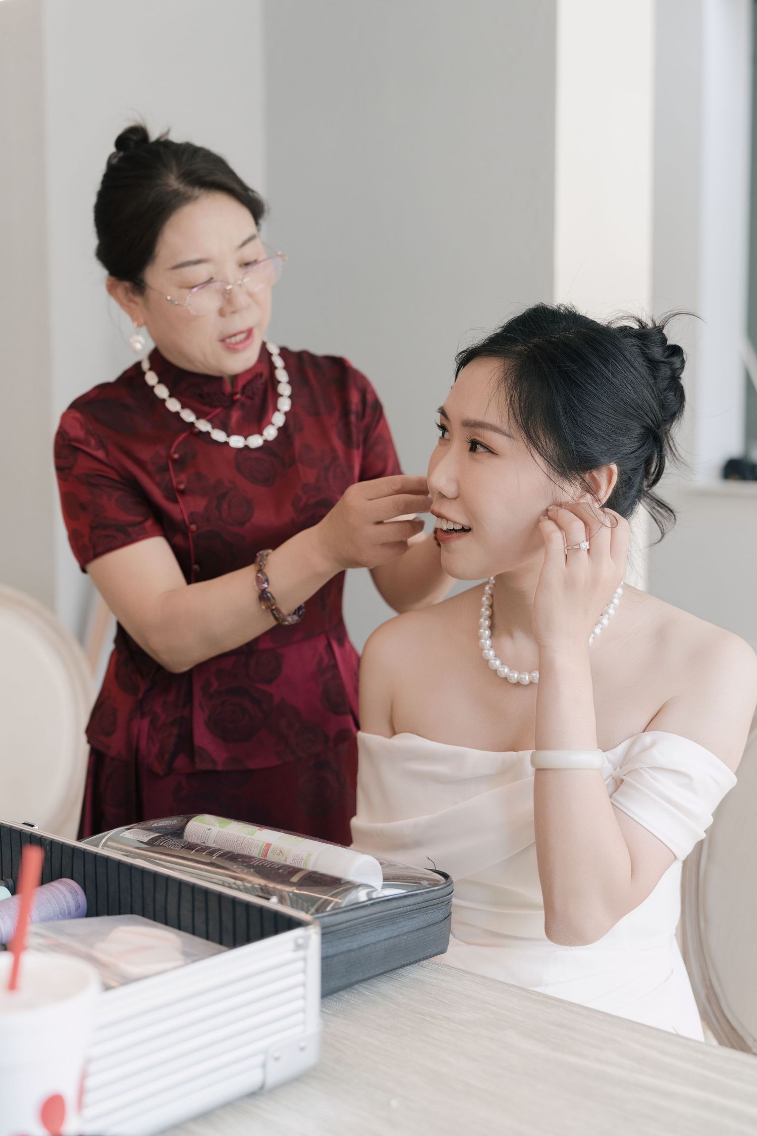 Woman in red qipao applies makeup to a smiling bride wearing a pearl necklace and white dress.