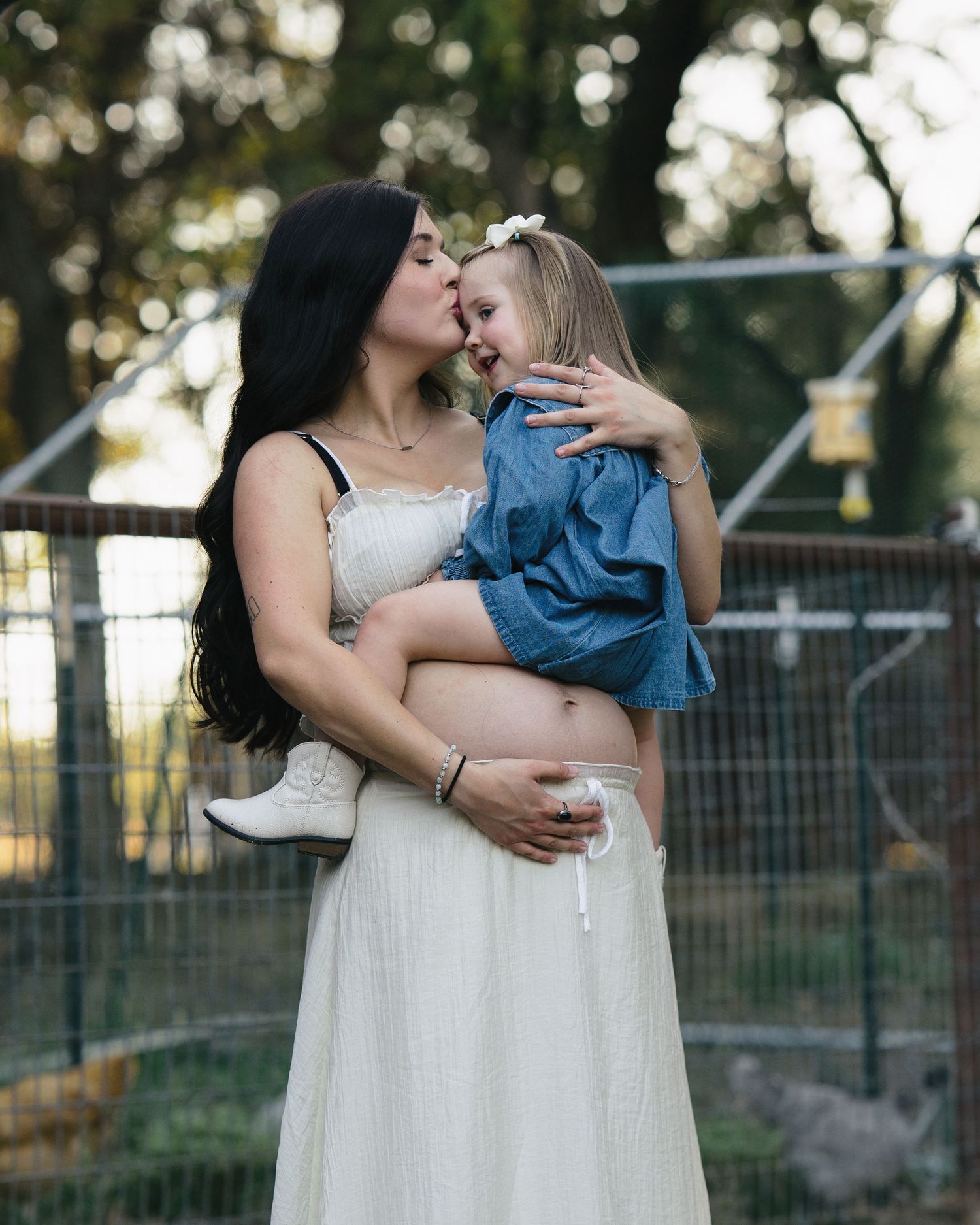 Pregnant mother kissing young blonde daughter while holding her at a farm during golden hour.