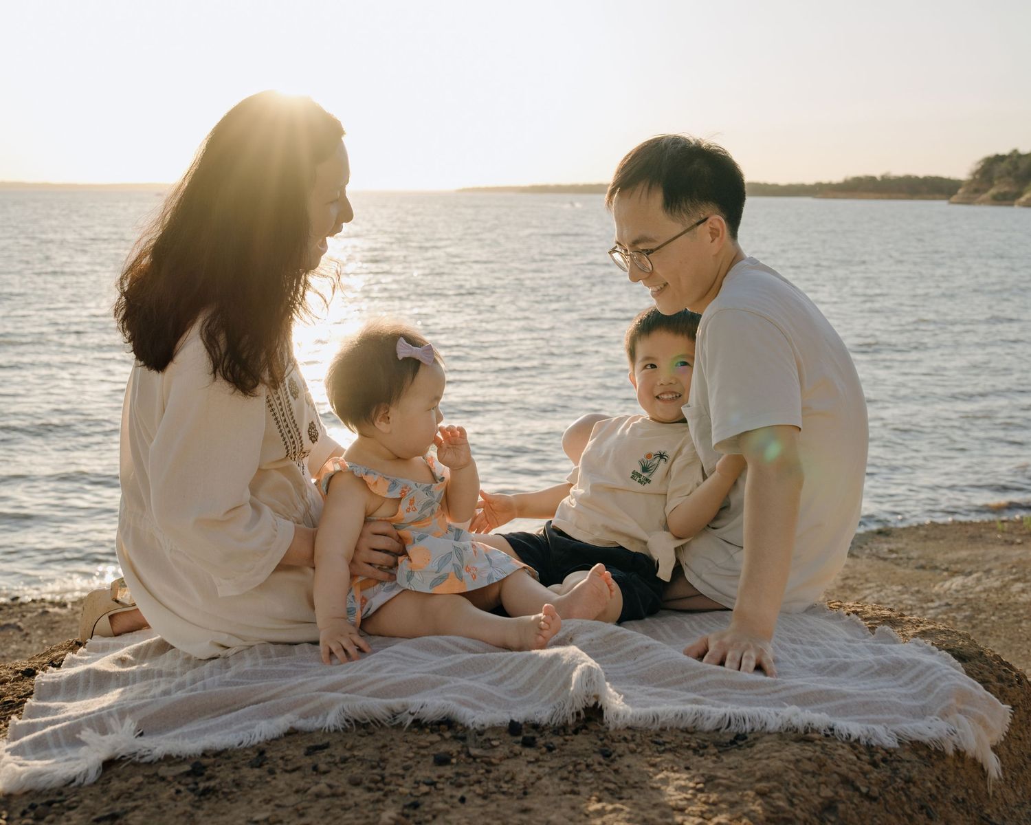 Family of four sits on blanket by the water at sunset enjoying a peaceful beach moment together.