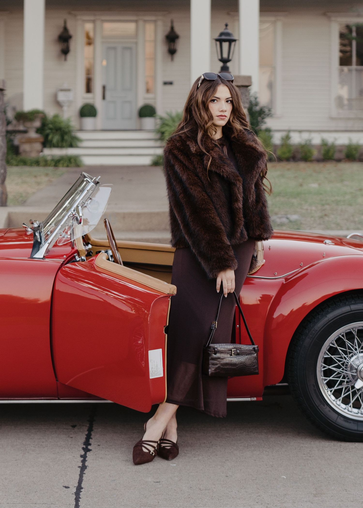 Stylish woman in fur coat holds crocodile handbag beside red vintage MGA convertible.
