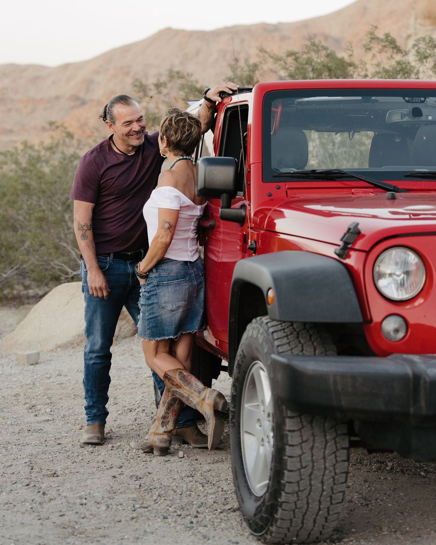 Man and woman sharing a moment next to a red Jeep Wrangler on a desert dirt road.