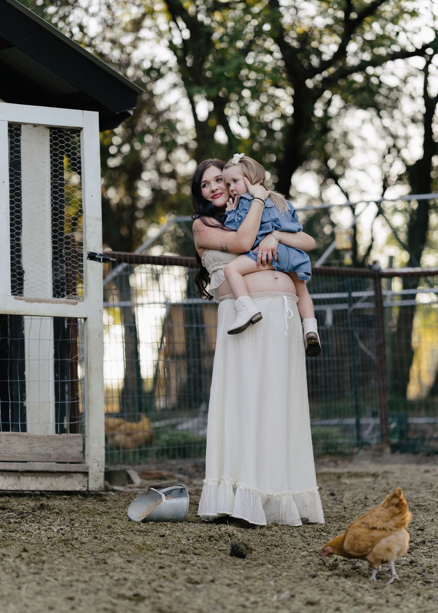 Pregnant woman in white dress holding toddler daughter near chicken coop on a rustic farm.