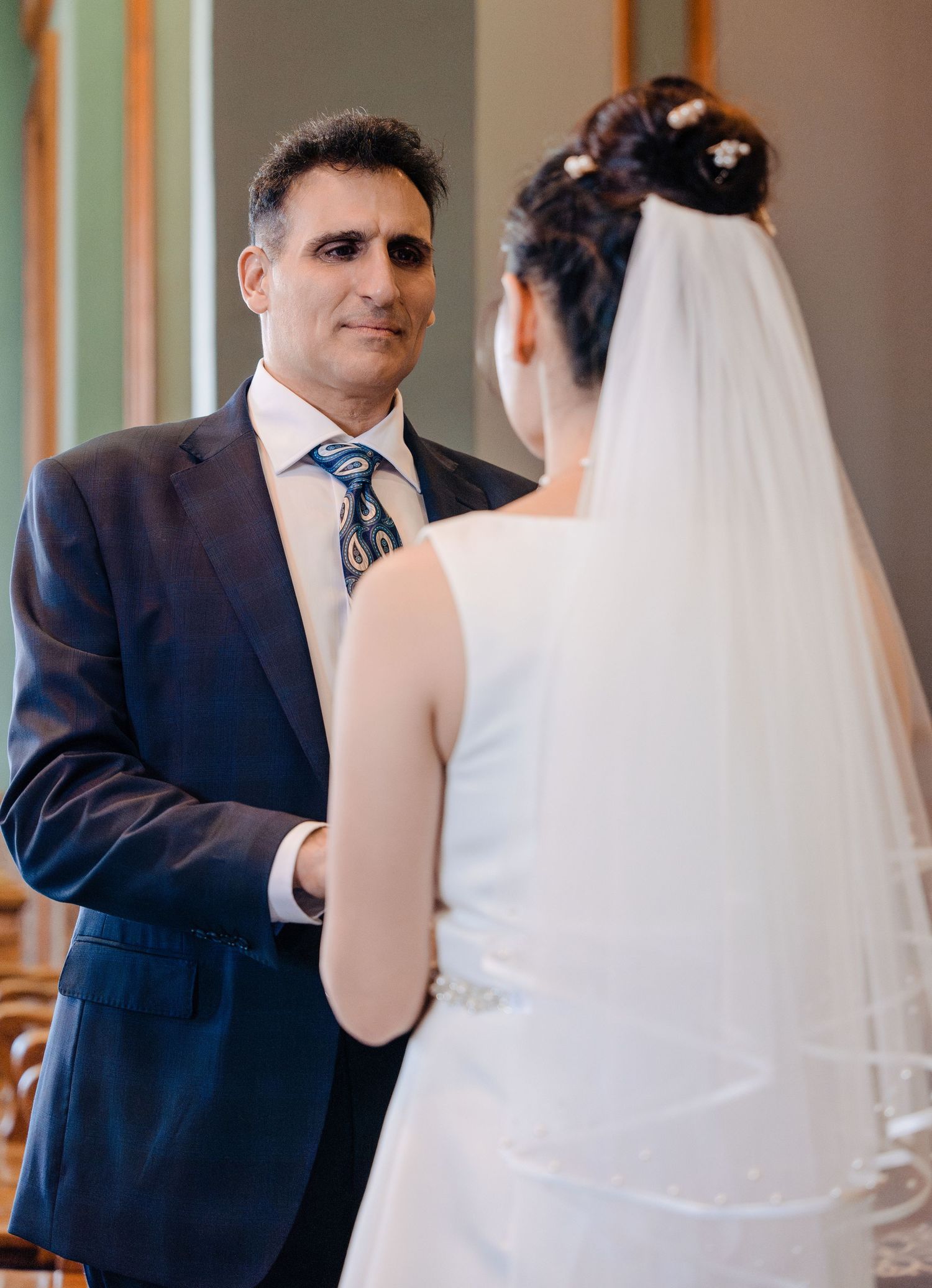 Groom in navy suit holds hands with bride in white wedding dress and veil indoors.