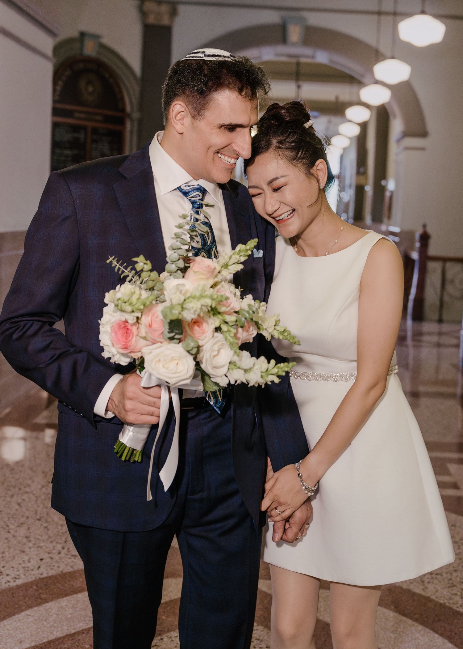 Smiling newlywed couple holding a pink and white bouquet inside a grand hall after their ceremony.