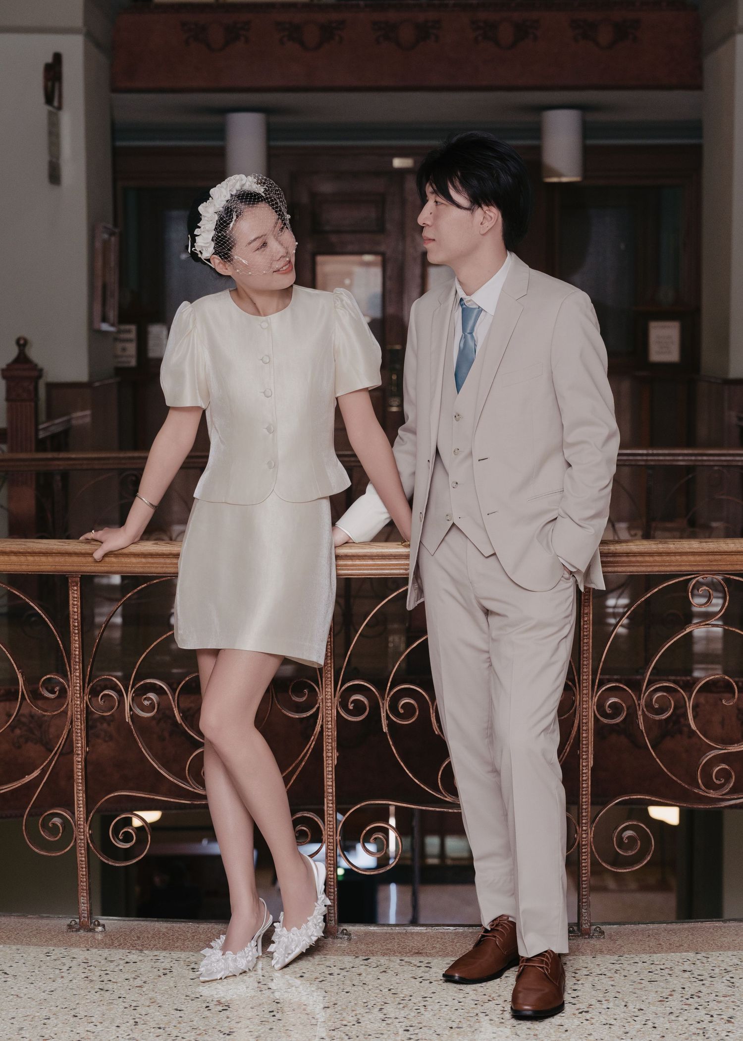 Bride in ivory mini dress and groom in beige suit share a tender moment by decorative iron railings.