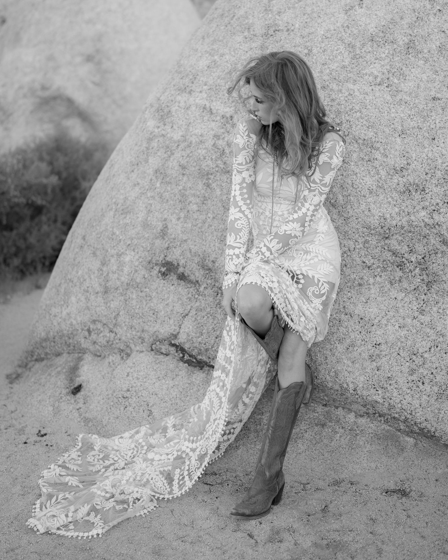 Black and white photo of bride in lace wedding dress and cowboy boots leaning against desert rock.