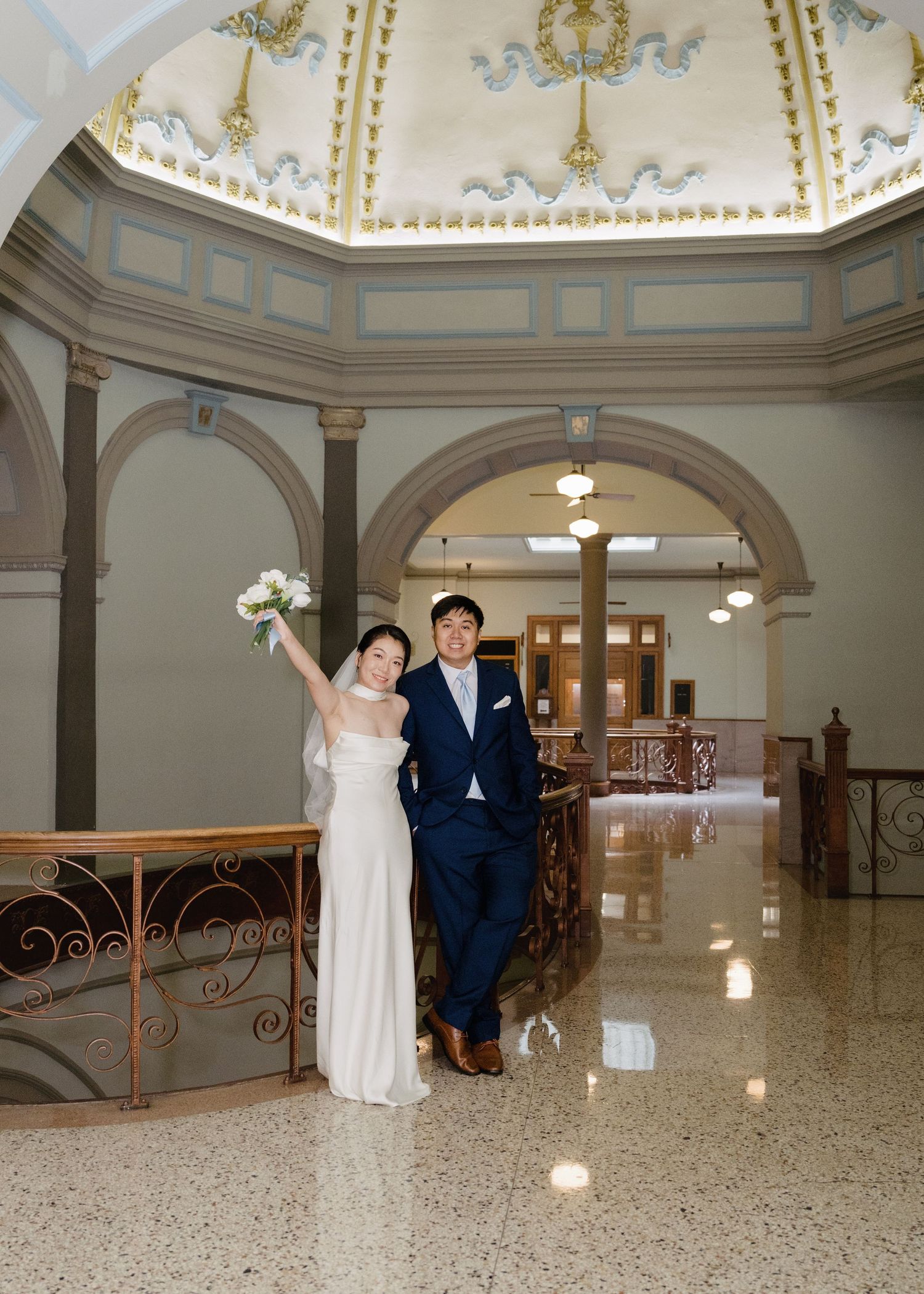 Newlywed couple posing in elegant wedding attire inside a grand hall with ornate domed ceiling and marble floors.