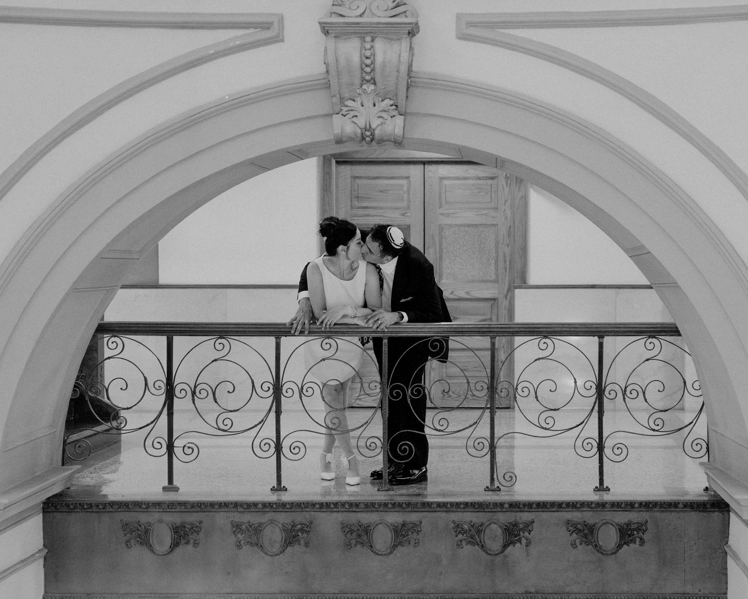 Black and white photo of a couple sharing a romantic moment on an ornate balcony with arched architecture.