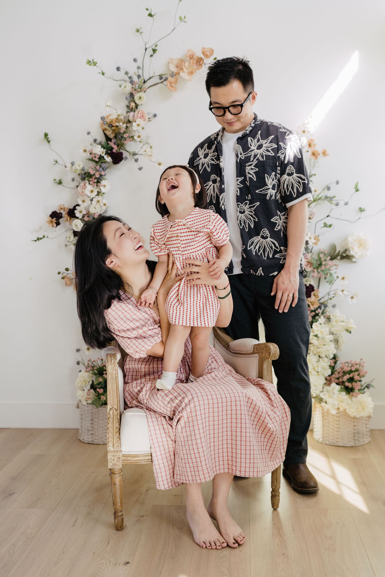 A joyful Asian family of three posing in front of a floral arch, mother and daughter in pink gingham dresses.