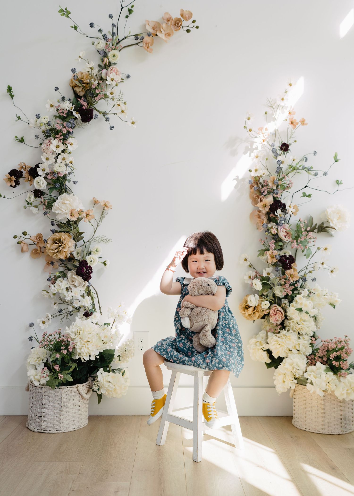 Smiling toddler girl in floral dress holding teddy bear on stool with flower arch backdrop.