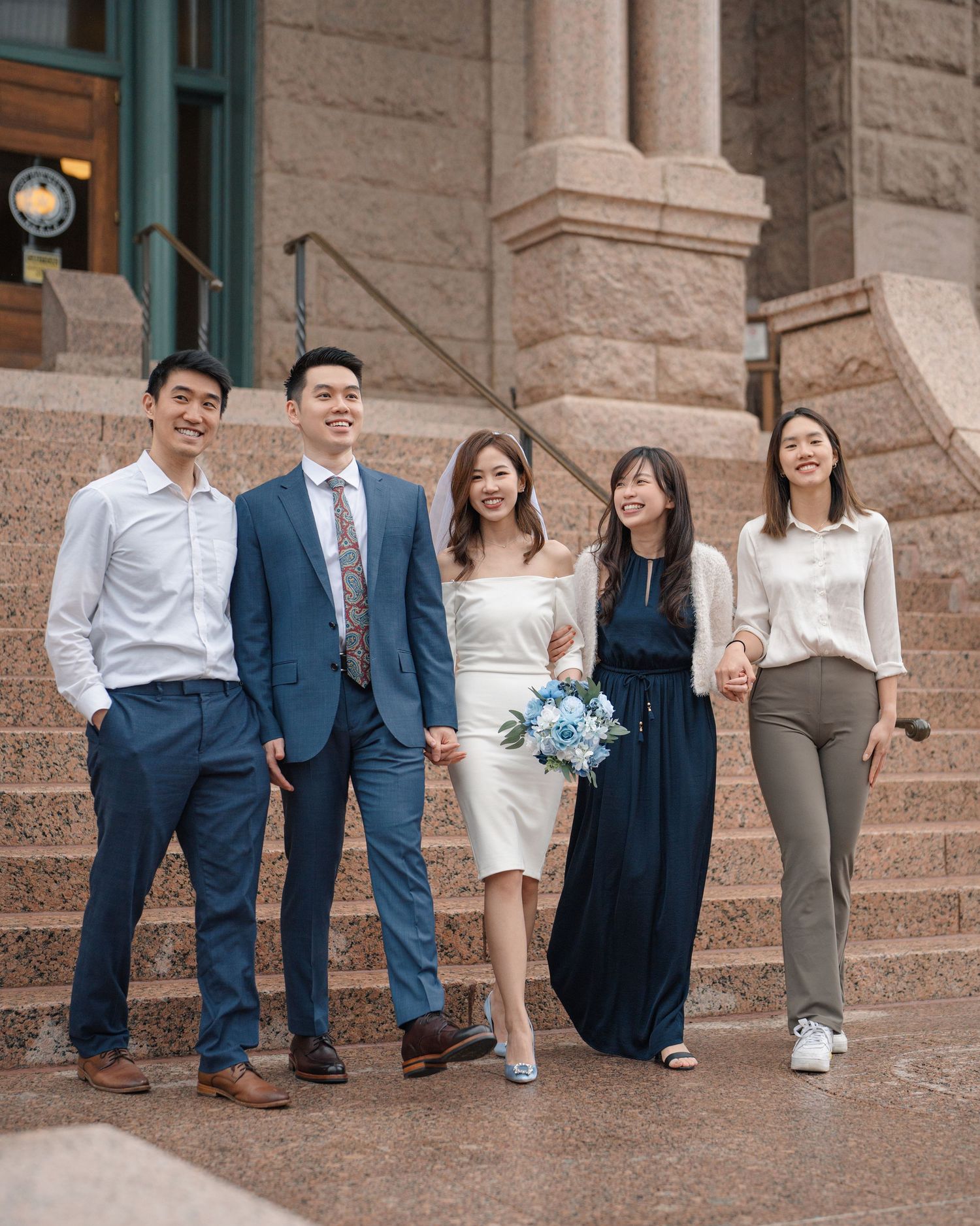 Asian wedding party posing on courthouse steps, bride holding blue and white floral bouquet.