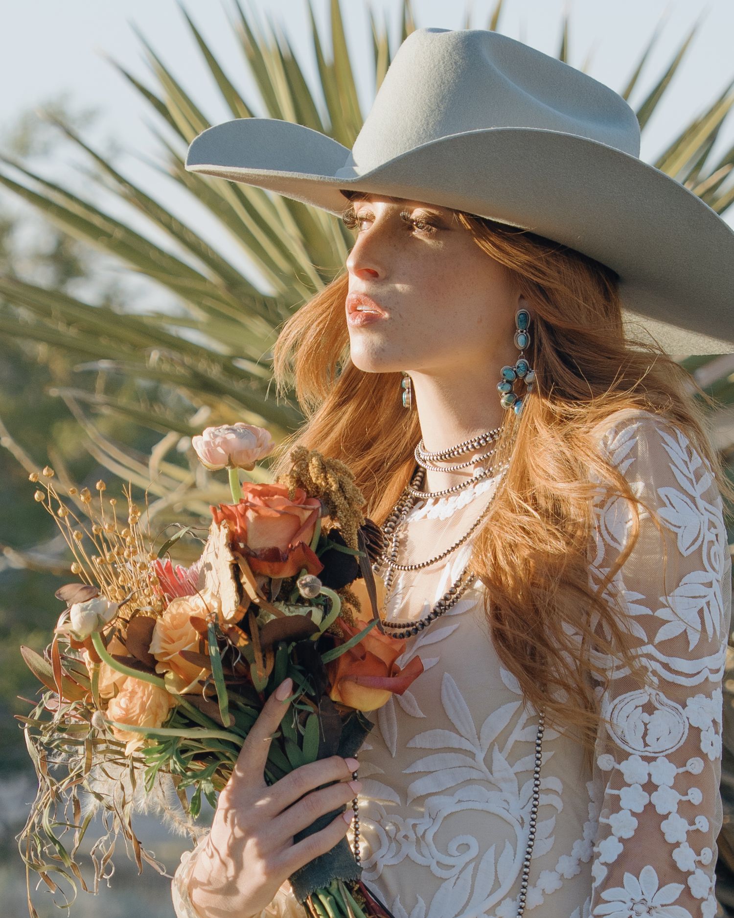 Boho bride in white lace dress and cowboy hat holding peach roses bouquet in desert setting.