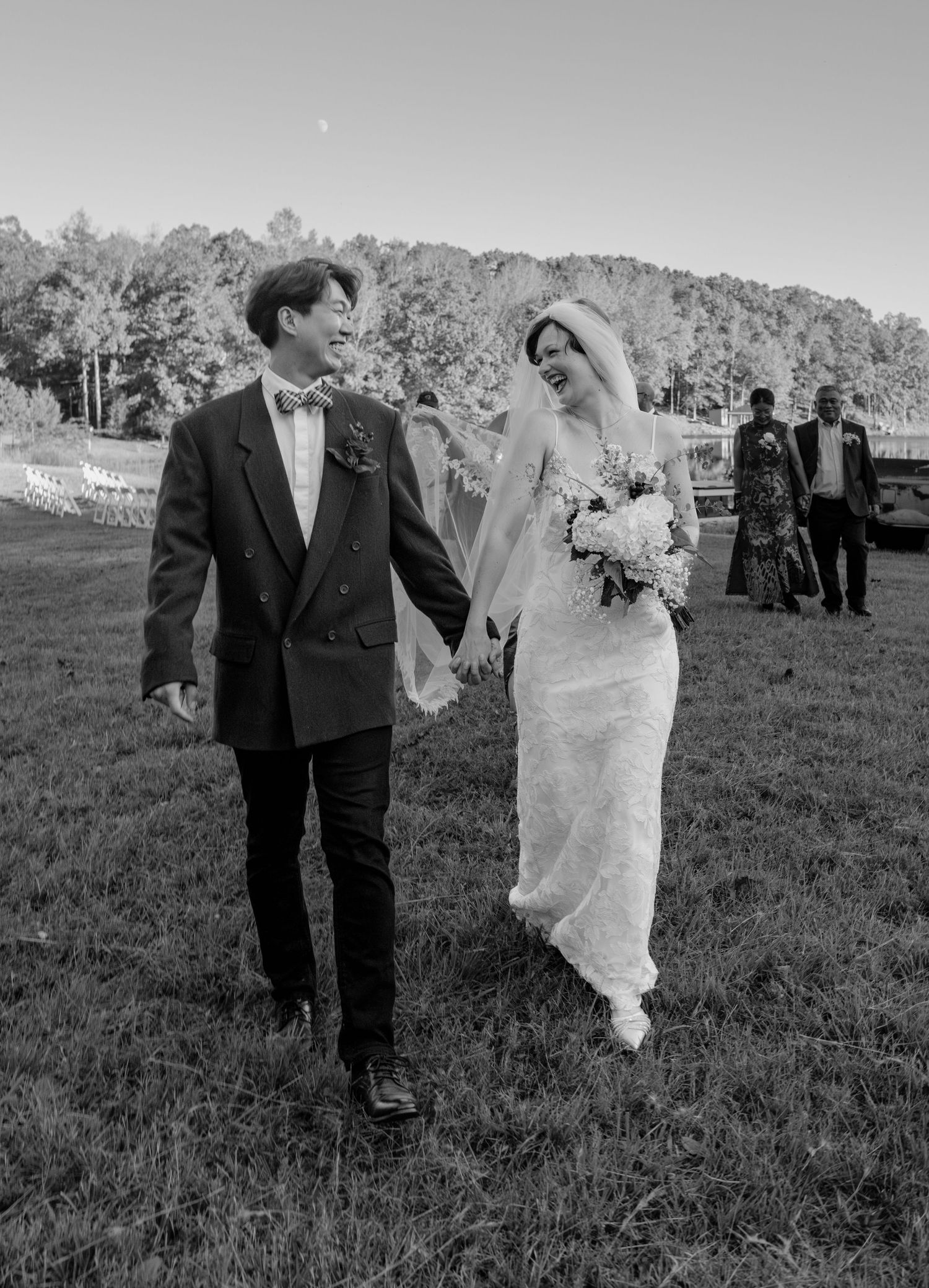 Black and white photo of happy newlyweds walking hand in hand across grassy outdoor venue.