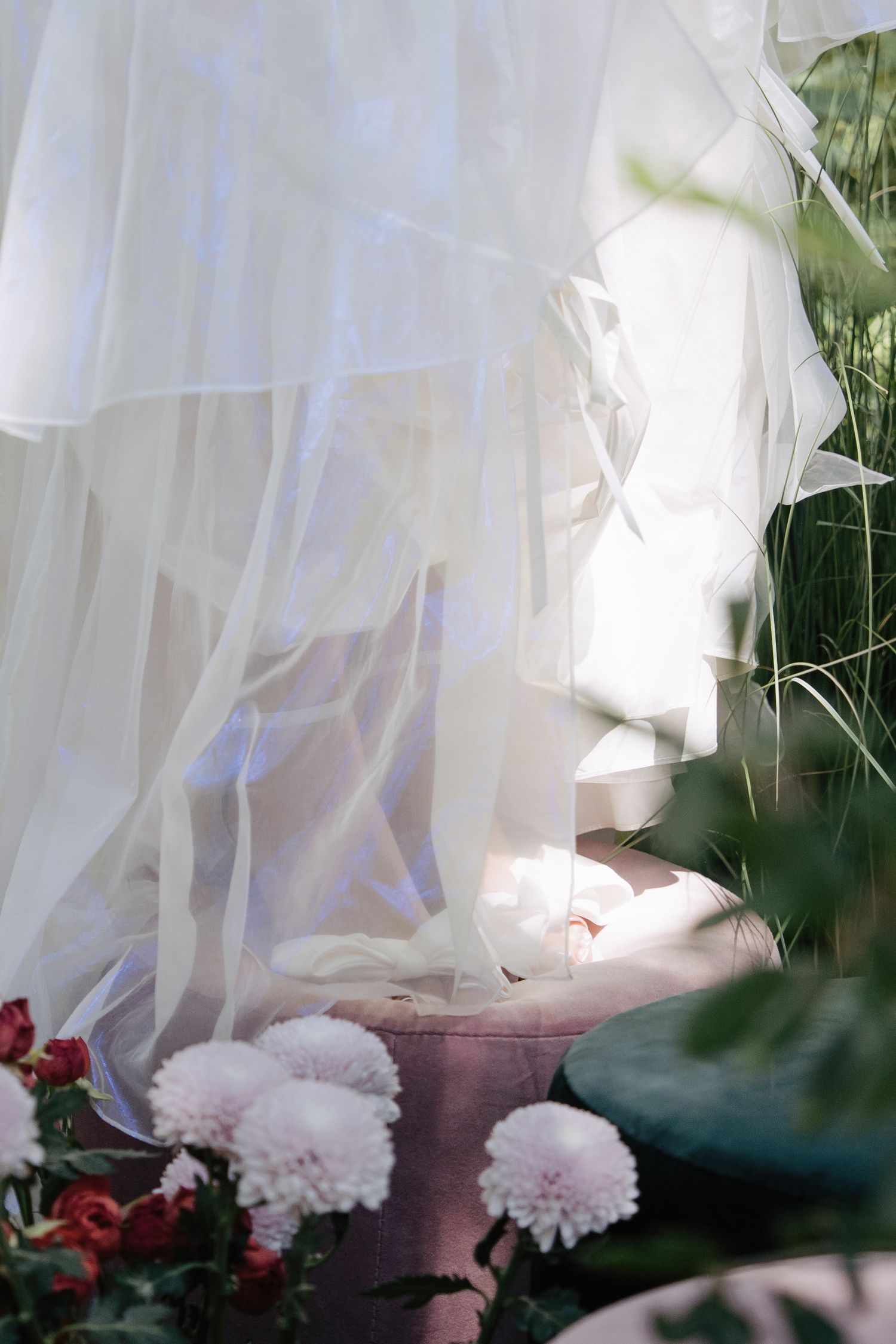 Flowing white bridal gown hem surrounded by pink chrysanthemums and lush greenery in soft natural light.