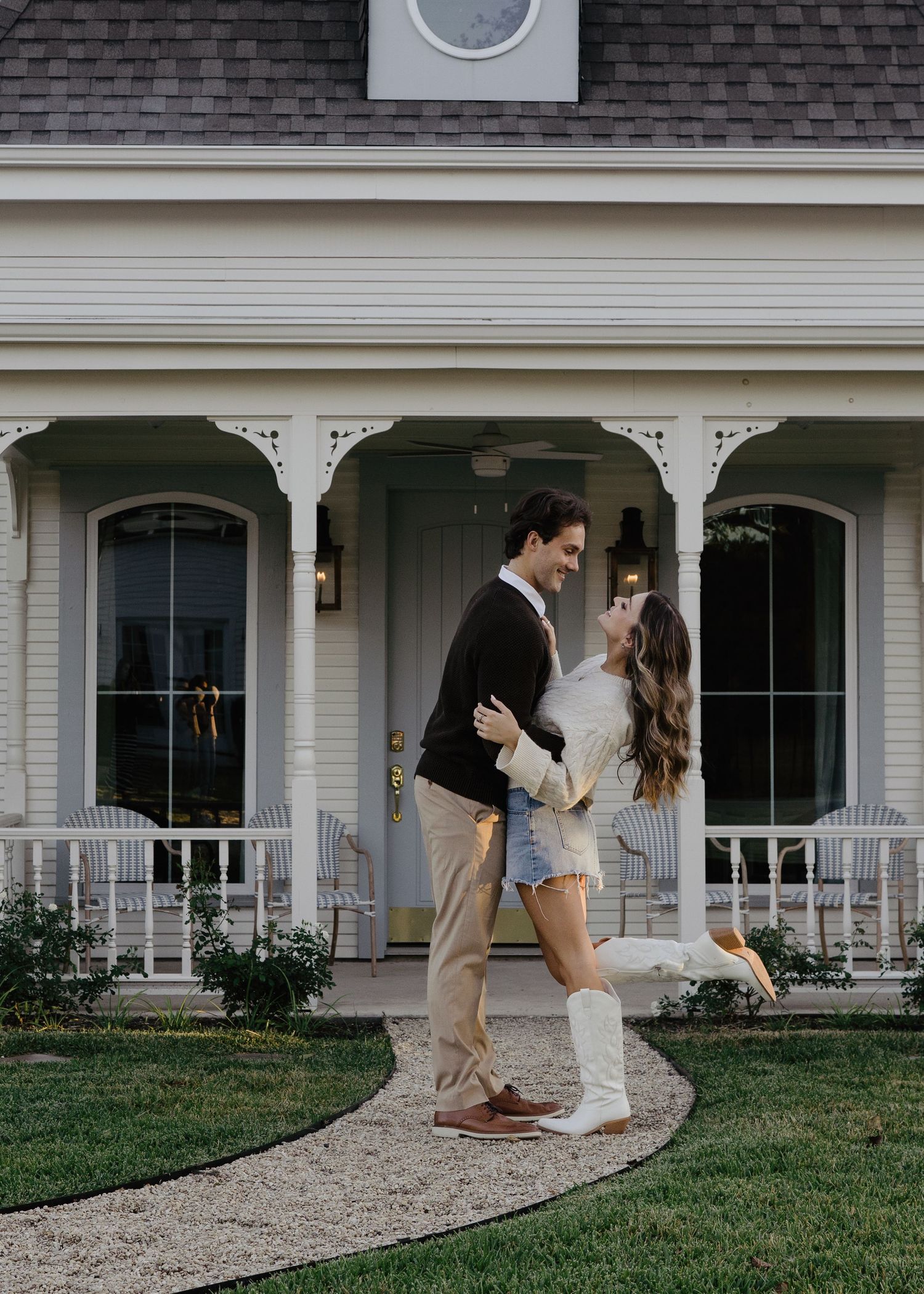 Couple embracing and smiling outside a charming white cottage with a covered front porch.