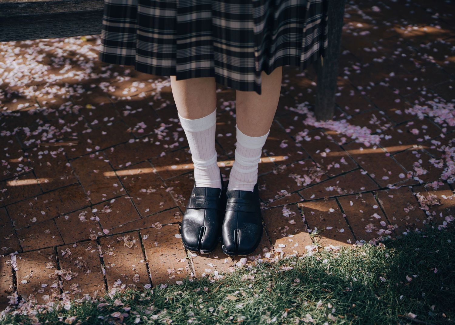 Close-up of legs in plaid skirt with white socks and black loafers on brick path with fallen petals.