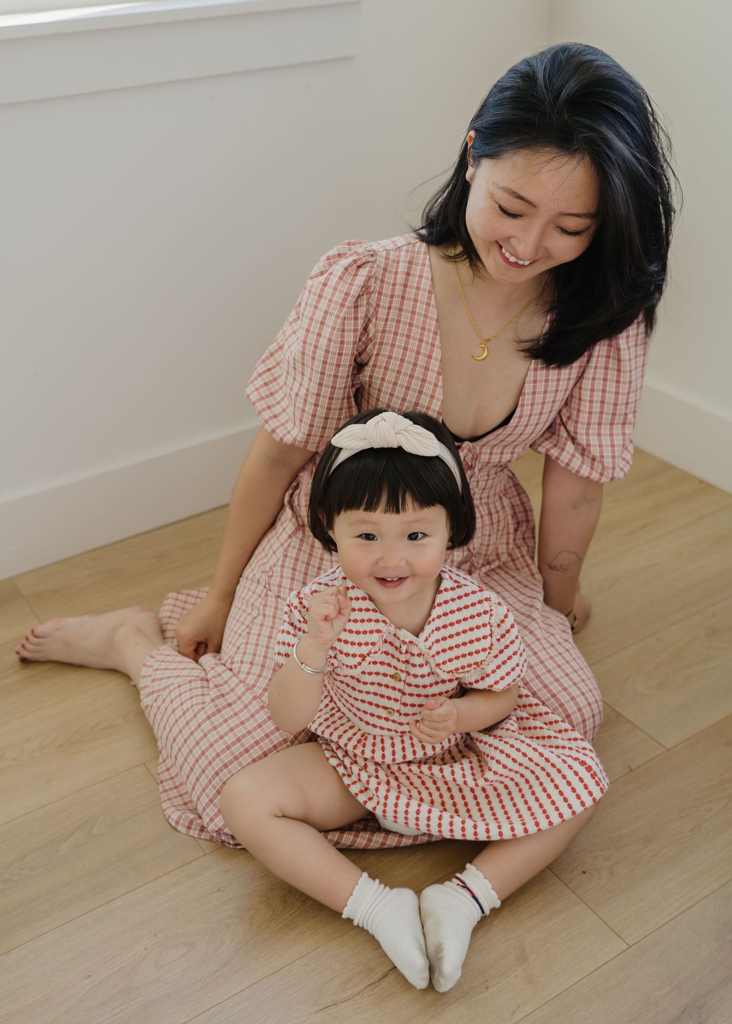 An Asian mother smiles warmly at her toddler daughter as they sit together on a light hardwood floor.