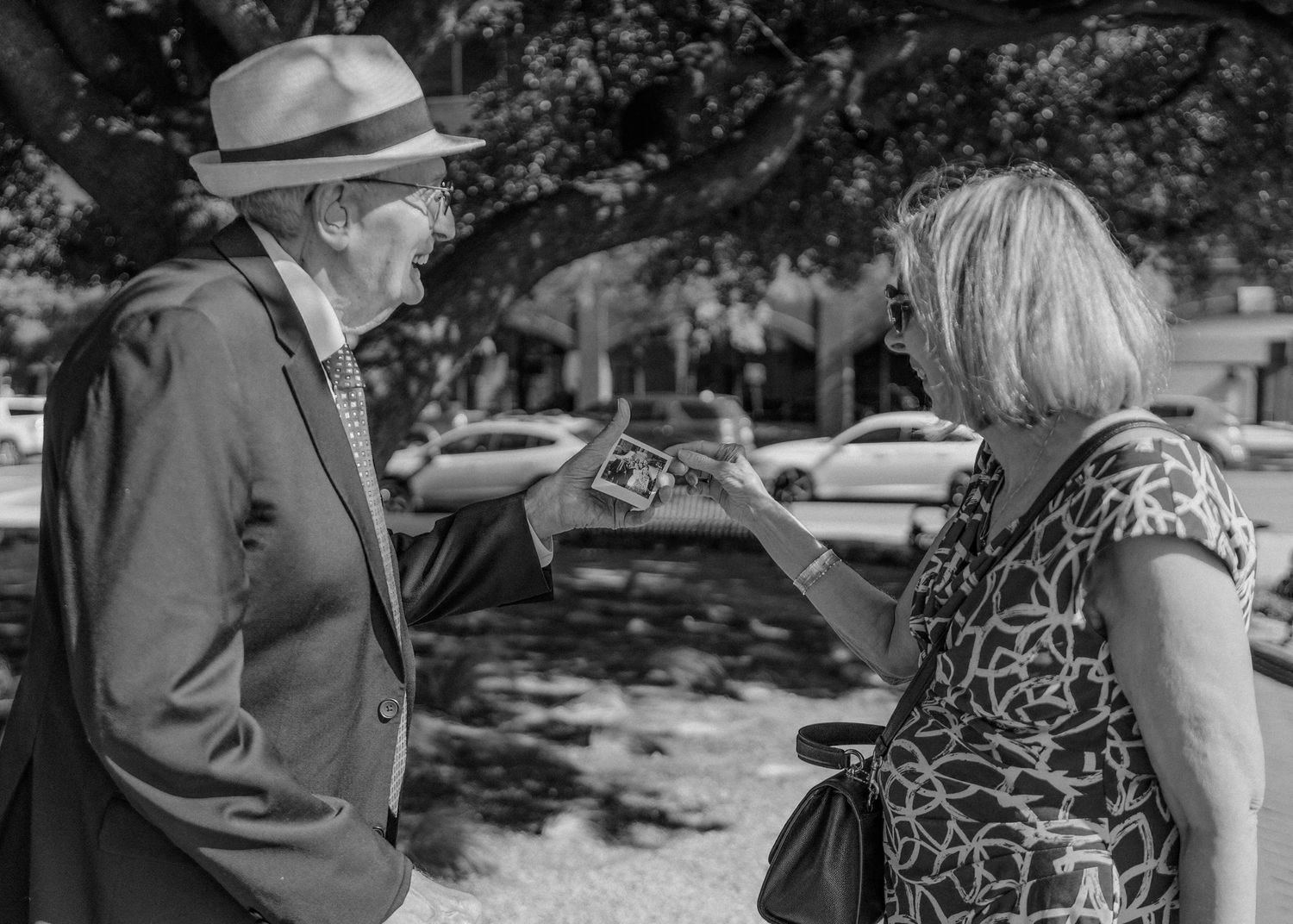 Elderly man in suit and hat converses with blonde woman outdoors at wedding reception.