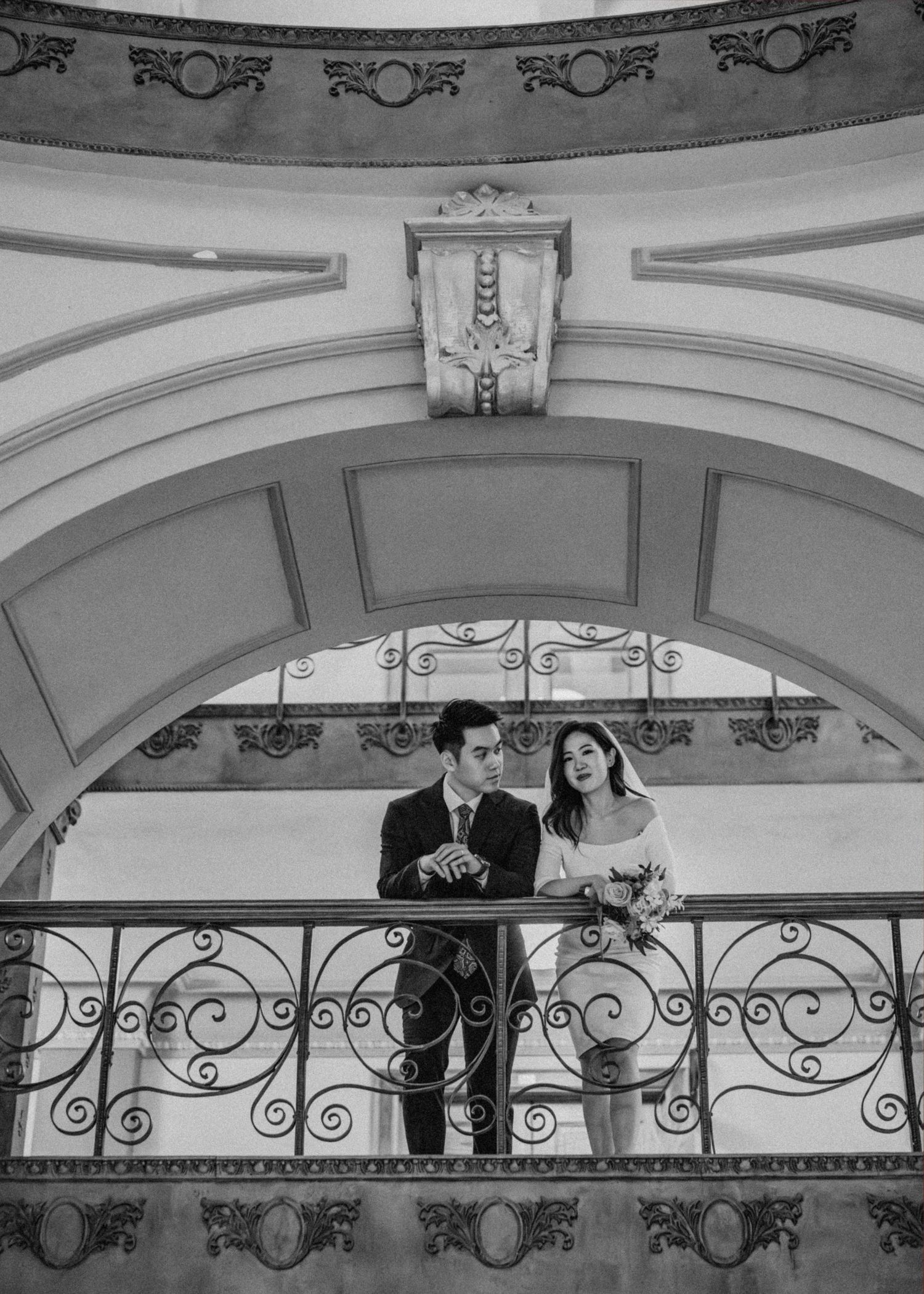 Black and white photo of newlyweds standing on decorative balcony under ornate arched ceiling.