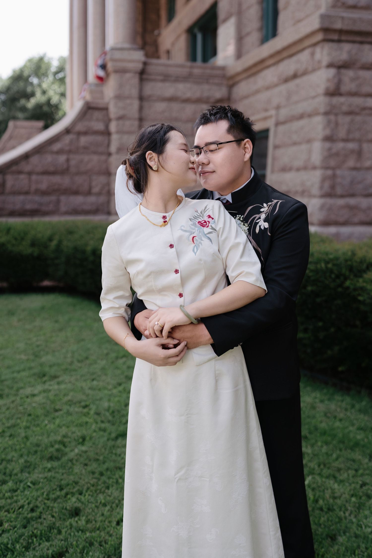 Couple in traditional Asian wedding attire embracing and sharing a kiss outside a historic stone building.
